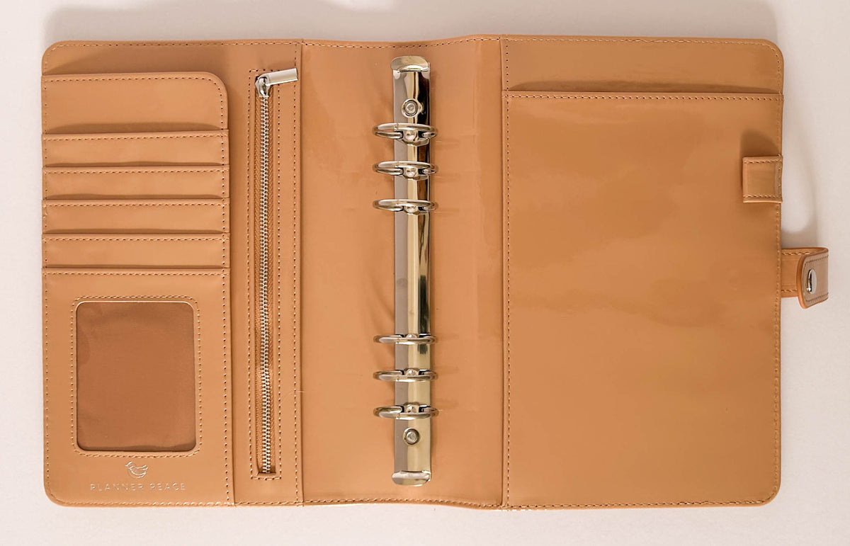 A tan leather ring binder planner cover shown open, featuring card slots, a clear ID window, a zippered pocket, and a central six-ring binder mechanism on a white background.