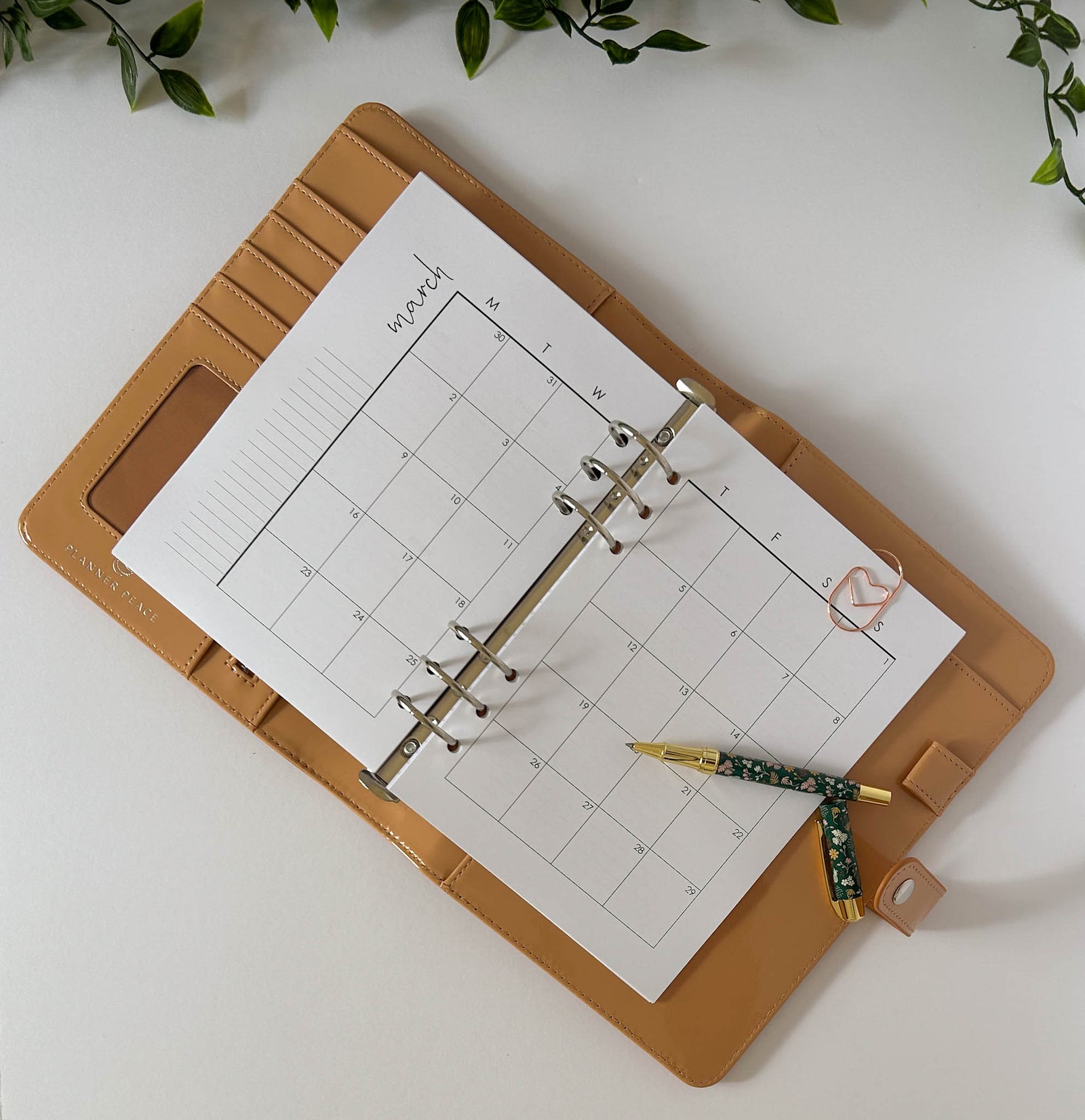 A tan leather planner binder with a weekly calendar page inserted, photographed from above on a white surface with green leaves at the top corners. A caramel bouquet pen rests on the planner.