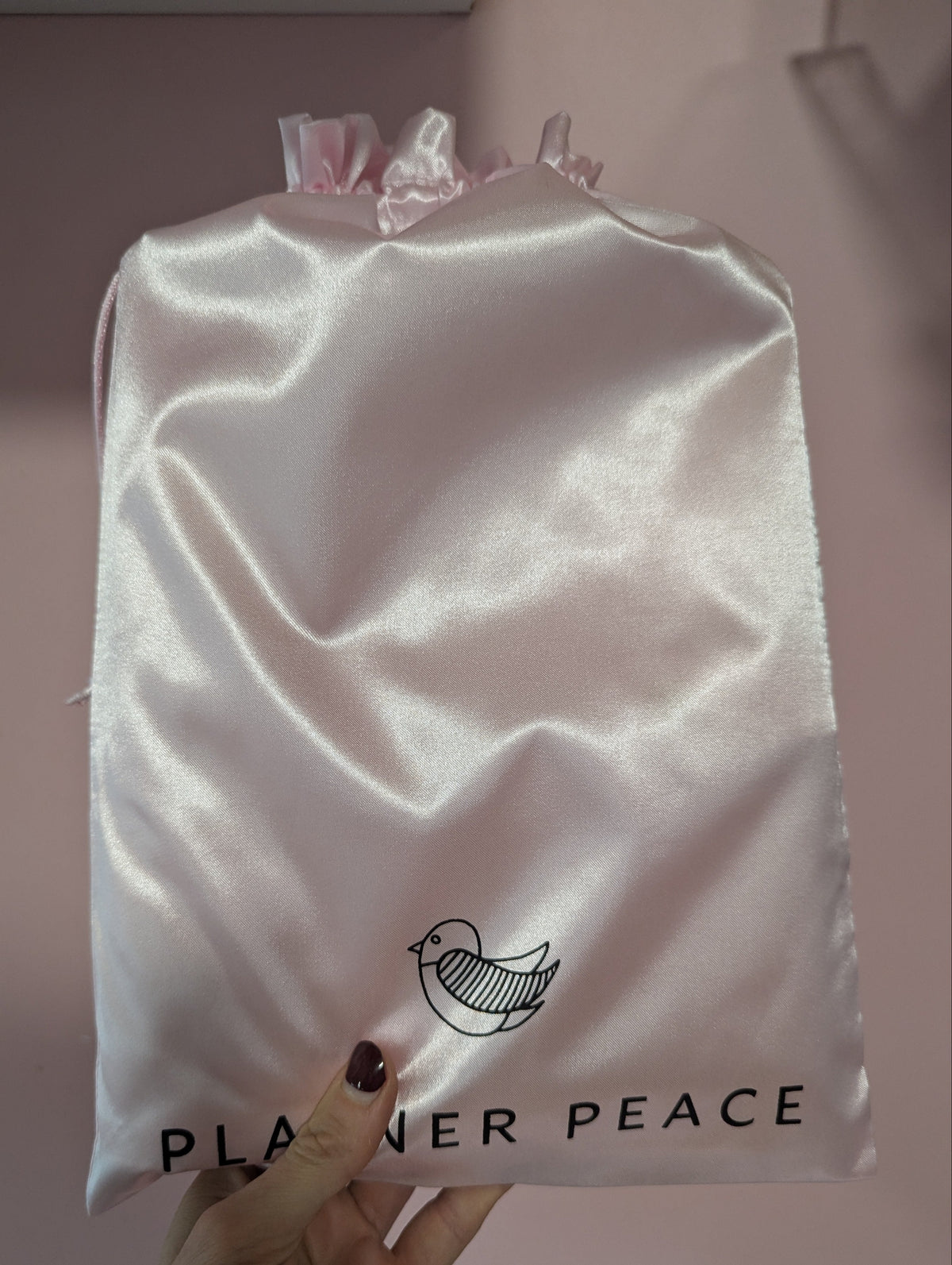White satin bag with &#39;Planner Peace&#39; logo held by a hand against a pink background